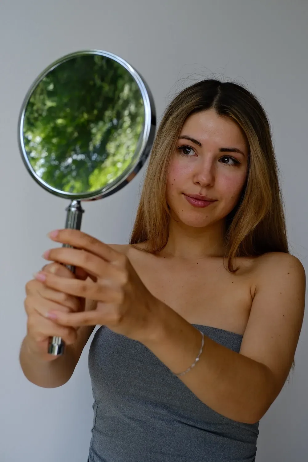 Frau mit langem Haar hält einen runden Handspiegel und betrachtet das Behandlungsergebnis ihrer Behandlung bei Skinthetics
