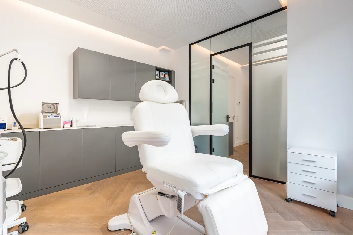 Behandlungsraum von innen:
Modernes kosmetisches Behandlungszimmer mit verstellbarem weißem Stuhl, grauen Schränken und Glas-Eingangstür.