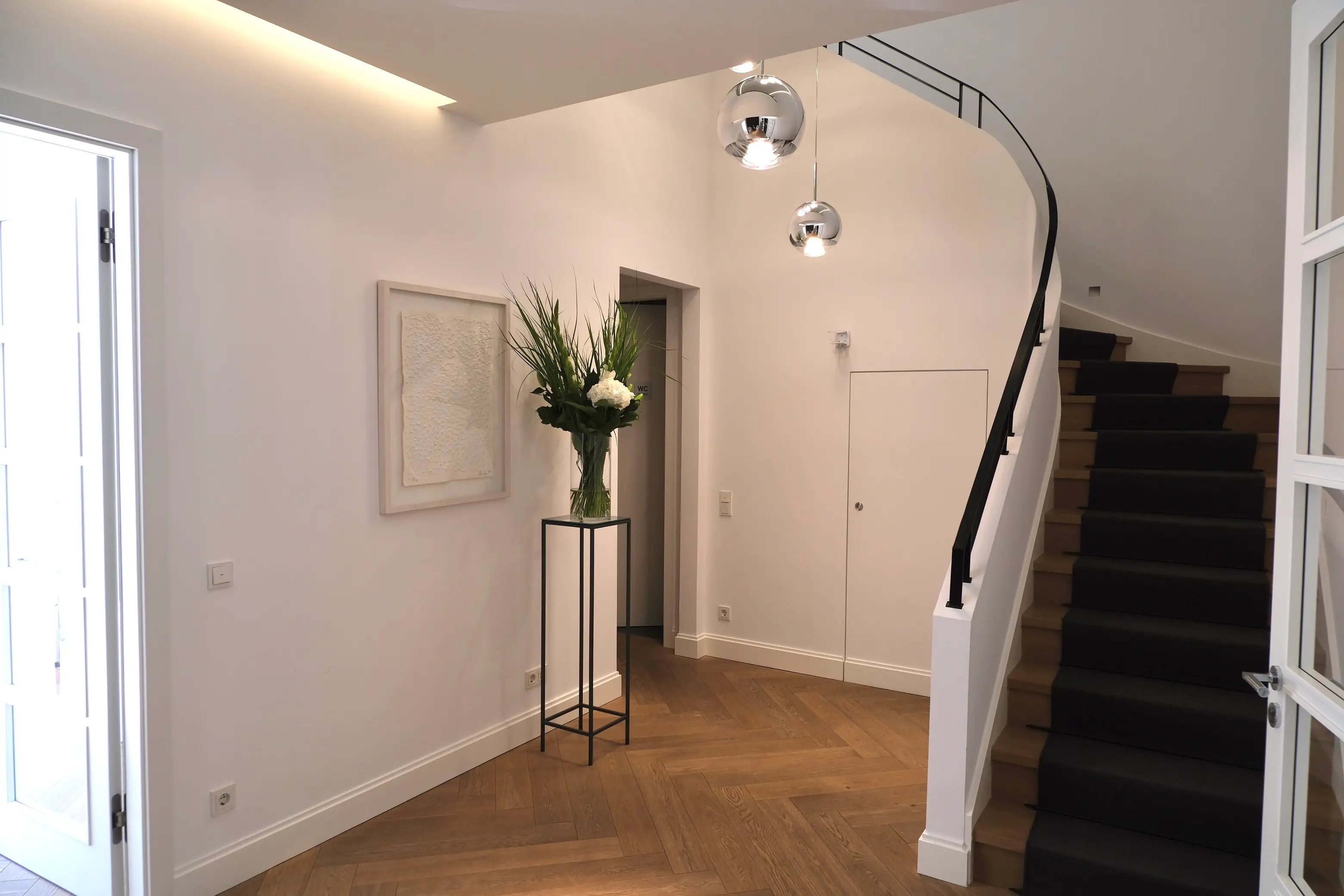 Kosmetik-Institut von innen:
Moderner heller Flur mit Holzparkett, Treppe mit schwarzem Teppichläufer, großem Blumenarrangement auf einem schmalen schwarzen Gestell und zwei hängenden Kugelleuchten.